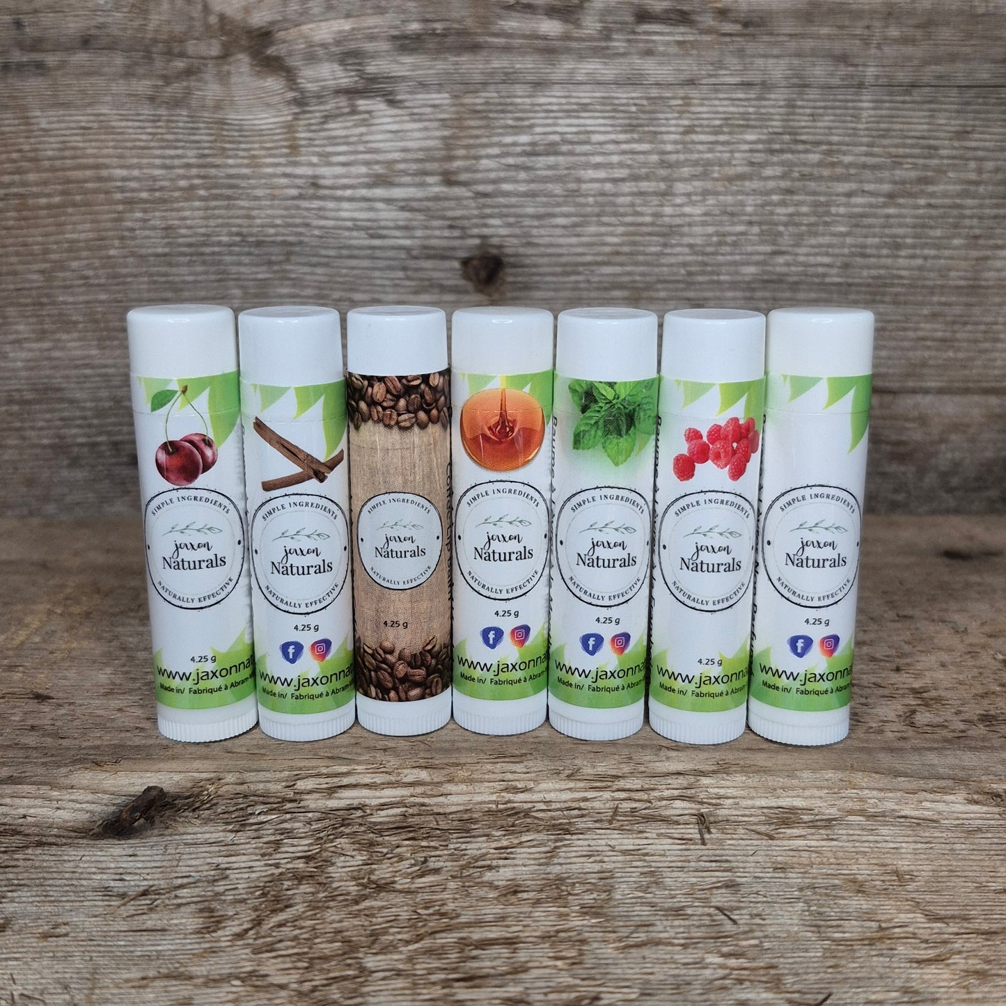 Seven handcrafted Jaxon Naturals lip balms in assorted flavours displayed on rustic barn wood, including coffee, cinnamon, maple, mint, raspberry, cherry, and unscented.