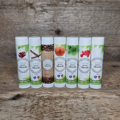 Seven handcrafted Jaxon Naturals lip balms in assorted flavours displayed on rustic barn wood, including coffee, cinnamon, maple, mint, raspberry, cherry, and unscented.