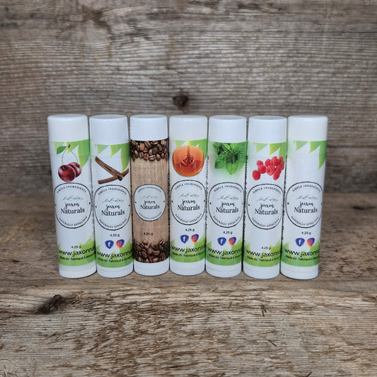 Seven handcrafted Jaxon Naturals lip balms in assorted flavours displayed on rustic barn wood, including coffee, cinnamon, maple, mint, raspberry, cherry, and unscented.