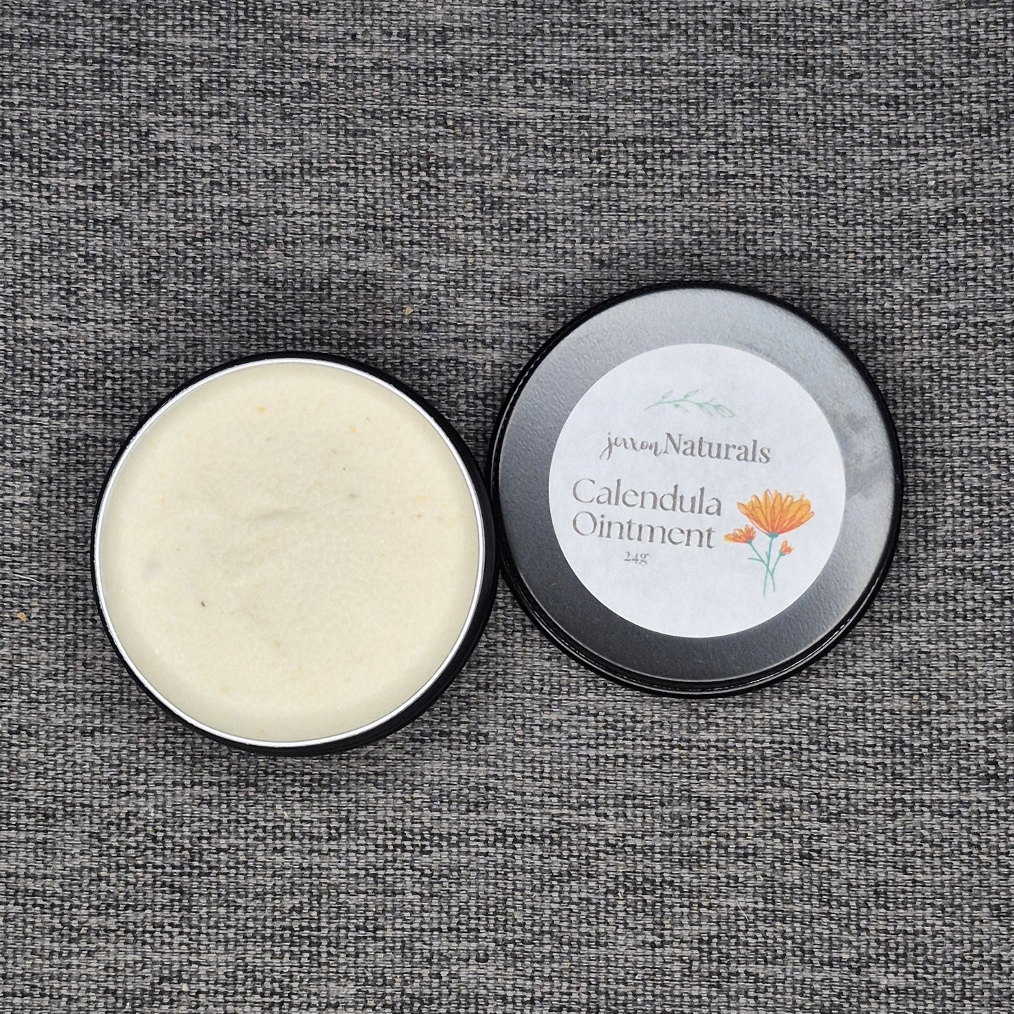 Open tin of Calendula Ointment with visible cream inside on a textured gray background